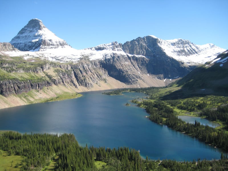 Snow capped alpine peak sits at the edge of a deep blue sub-alpine lake in Glacier National Park.