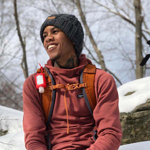 Justin Tucker is geared up with a warm hat, jacket, and backpack for a wintery portrait with snow and bare trees in the background.