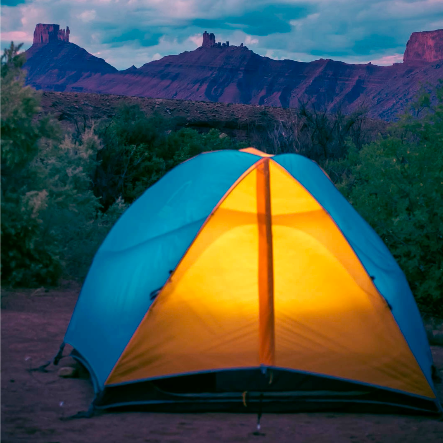 A yellow and blue tent glows in front of a pink and purple lit canyon in the background.