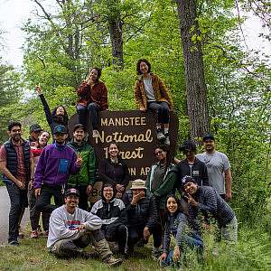 A group of Black and Brown Chicagoans gather for a group photo in front of the Huron-Manistee National Forest sign in Michigan.