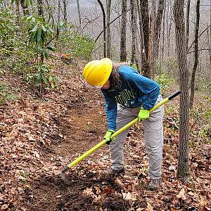 A volunteer in a yellow hardhat and neon gloves uses a hoe to clean leaves and debris from a section of trail.