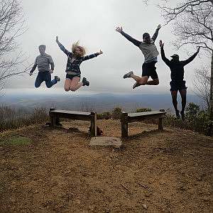 Four student volunteers jump in unison from benches at a mountain overlook with fog and rolling hills in the background.