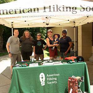 Five American Hiking Society staff and board members and one small child stand under a canopy tent that reads "American Hiking Society" with a table of info in front of them covered in a green tablecloth that also reads "American Hiking Society" with the AHS logo shaped like the bottom of a hiking boot.