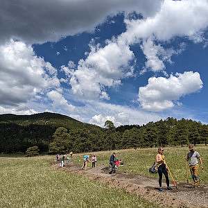 A group of volunteers work along a trail in a grassy meadow with a forested hill and white puffy clouds in the background.