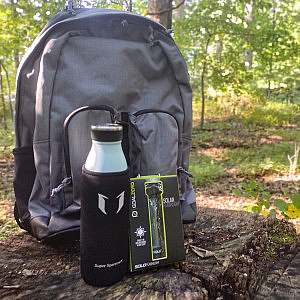 A water bottle and flashlight its packaging site in front of a gray backpack which sits on a tree stump in a forest with light peeking through the leaves.