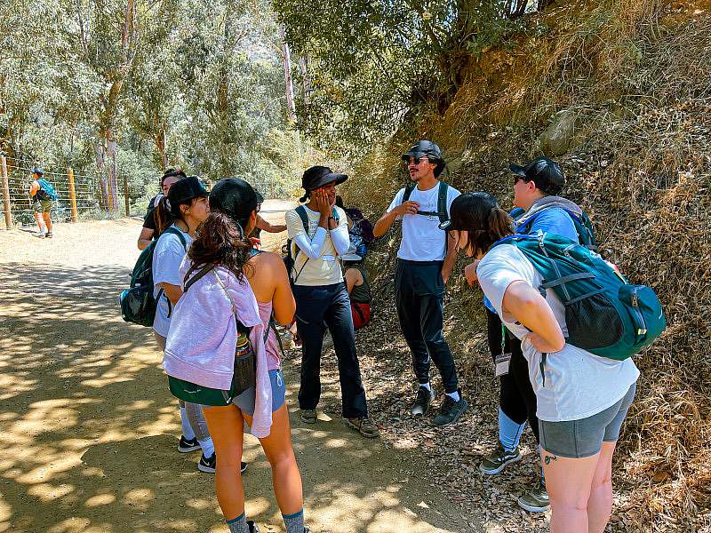 A diverse group of participants smile while they circle up in a shady section of the trail