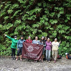 A group of volunteers stand together in front of tall evergreens holding a Latino Outdoors banner.