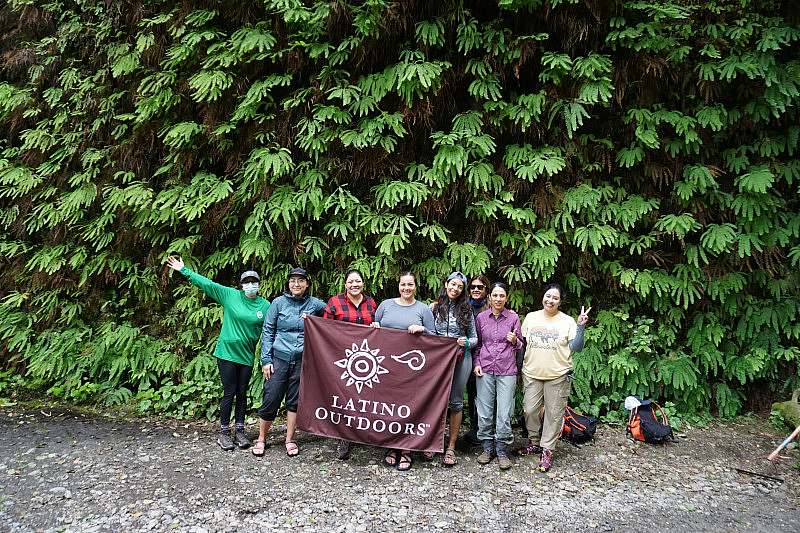 A group of volunteers stand together in front of tall evergreens holding a Latino Outdoors banner.