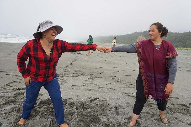 Two ladies hold hands dancing on a sandy beach with surf and forest in the background