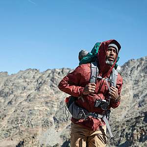 Hiker holds their pack straps as they hike with a warm hat and jacket with a hood with a blue sky and rugged ridge in the background.
