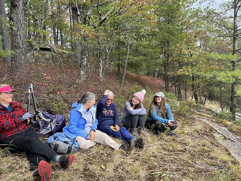 Five AHS trail volunteers sit near their worksite talking and laughing with forest & trees behind them