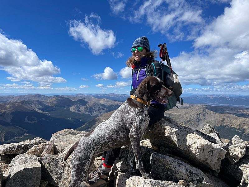 Hike with a dog stands on a rocky summit wearing a warm hatt with blue sky and puffy clouds and mountains in the background
