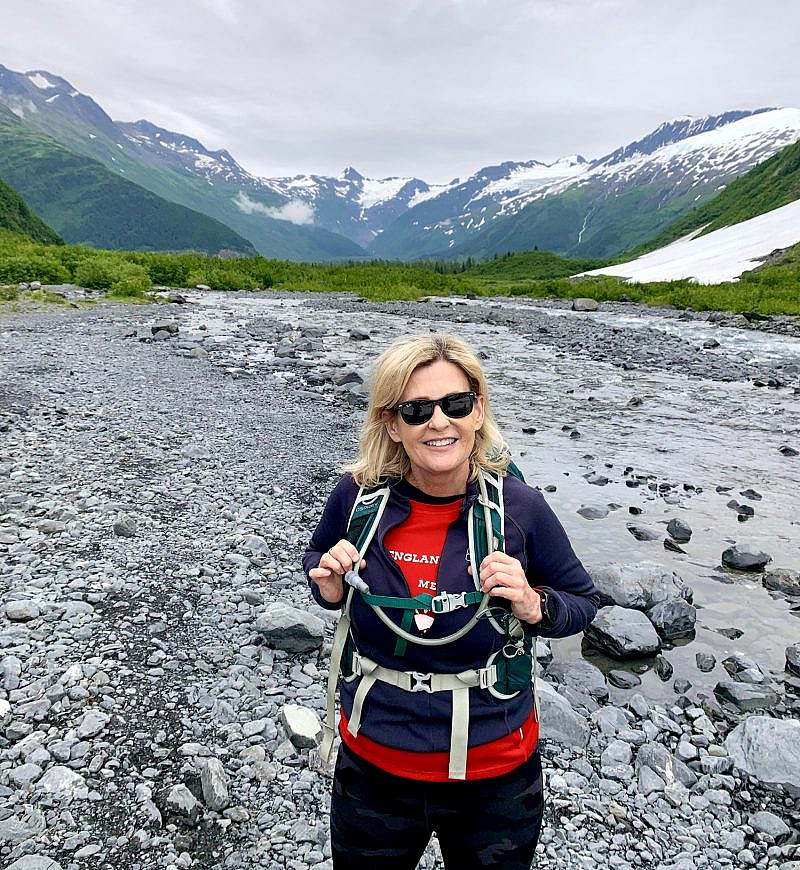 Hiker wearing sunglasses and holding both backpack shoulder straps stands in a rocky vally with snowy mountains in the background