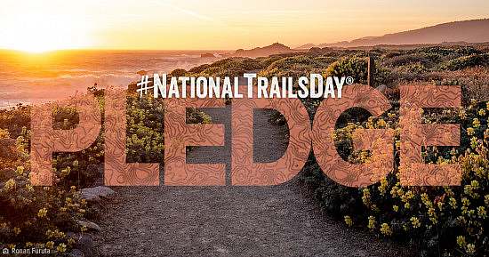 American Hiking Society's National Trails Day Pledge wordmark over lay on gravel trail winding through flowering bushes near a coastline as the sun sets.