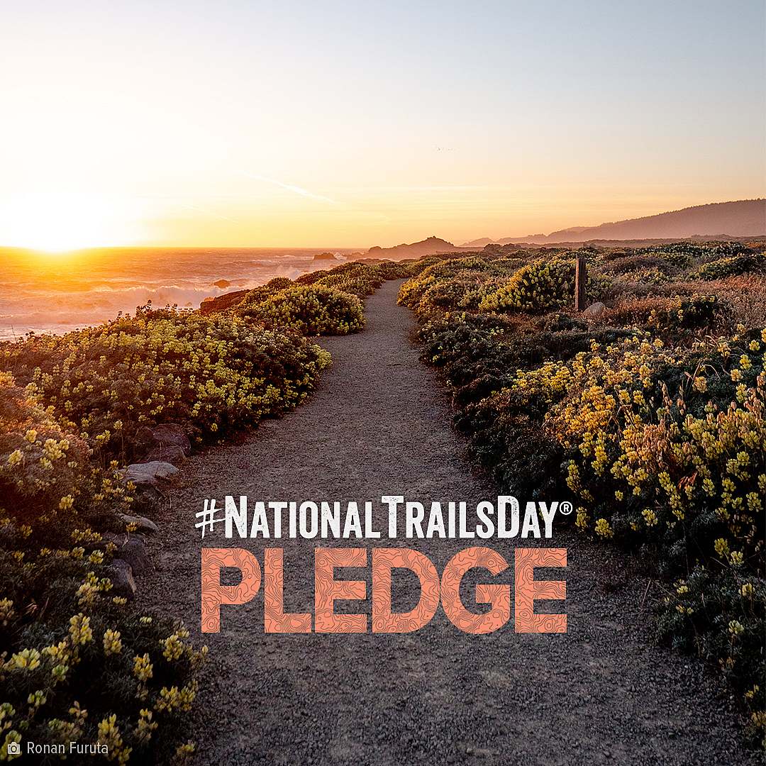 American Hiking Society's National Trails Day Pledge wordmark over lay on gravel trail winding through flowering bushes near a coastline as the sun sets.