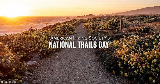 American Hiking Society's National Trails Day wordmark over lay on gravel trail winding through flowering bushes near a coastline as the sun sets.