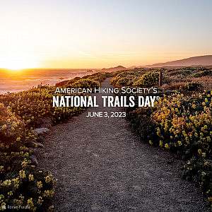 American Hiking Society's National Trails Day wordmark over lay on gravel trail winding through flowering bushes near a coastline as the sun sets.