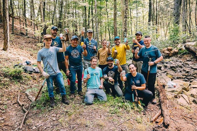 A group of volunteers wearing Athletic Brewing Co and Two for the Trails shirts hold trail tools in one hand as they lift up cans of Athletic Brewing for a group photo. 