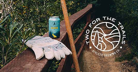 Two for the Trails logo overlaid on a photo of a can of Athletic Brewing Co. siting on a trail railing with work gloves and a wooden tool handle