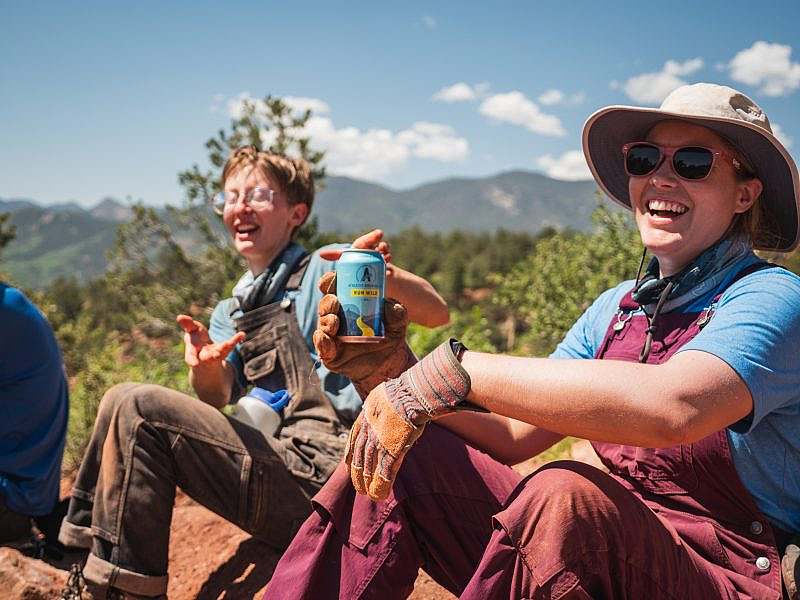 Two people in overalls laugh while taking a break on the side of the trail with a blue can of Athletic Brewing Co.
