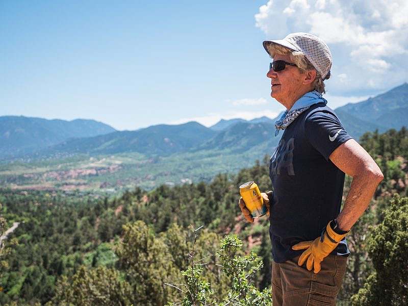 Person wearing work gloves and a bandana holds a yellow can of Athletic Brewing while gazing out across a wide landscape of trees and distant hills.