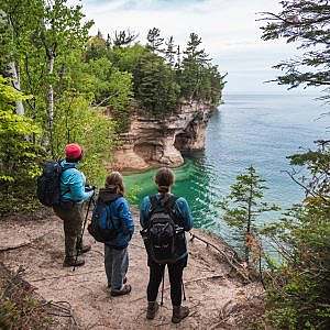 Three hikes gaze at a large body of water from the tree line atop cliffs above the water.