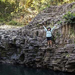 A hikerin in black shorts and a light long sleeve shirt stands with arms lifted high to celebrate stands on a cliff edge above a body of water.
