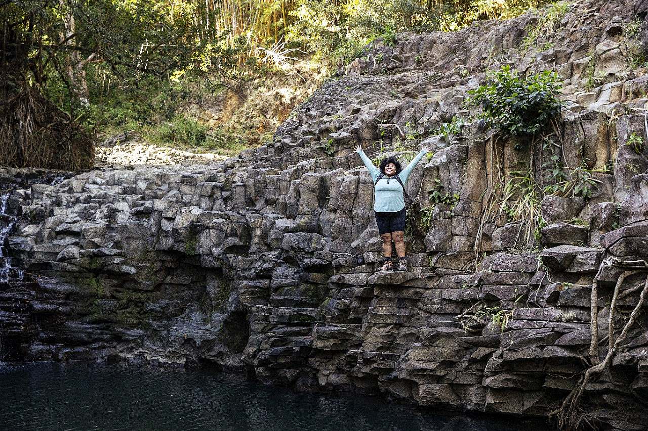 A hikerin in black shorts and a light long sleeve shirt stands with arms lifted high to celebrate stands on a cliff edge above a body of water.