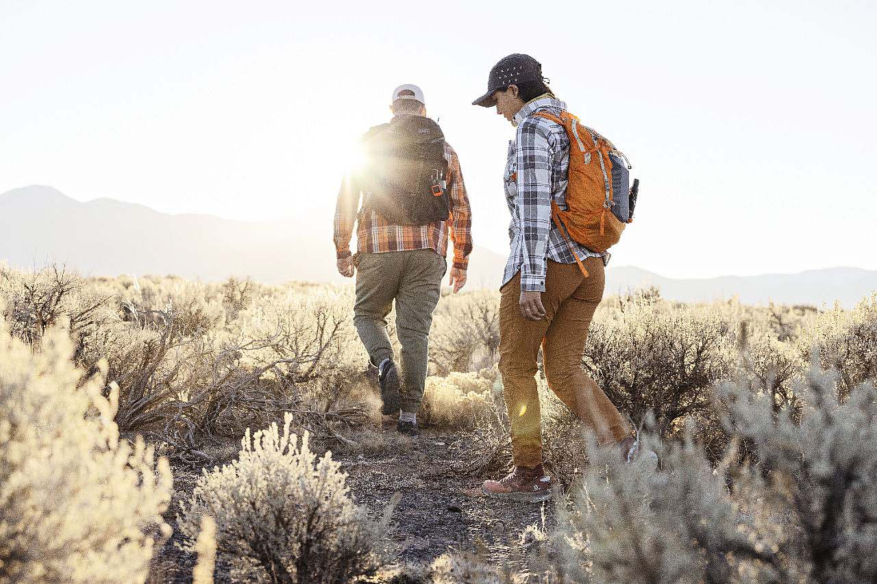 two hikers walk through sage brush as the sun rises on the horizon.