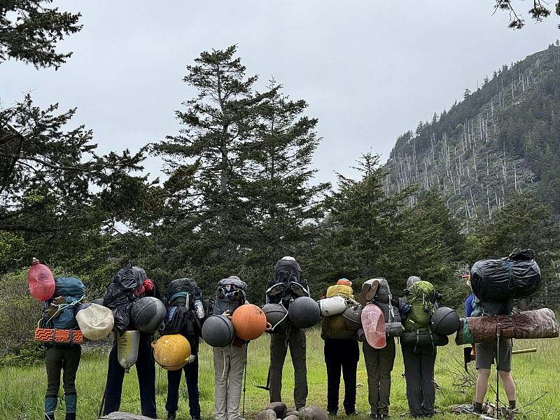 Nine people stand with their backs to the camera, showcasing their full backpacks with pieces of marine debris attached.