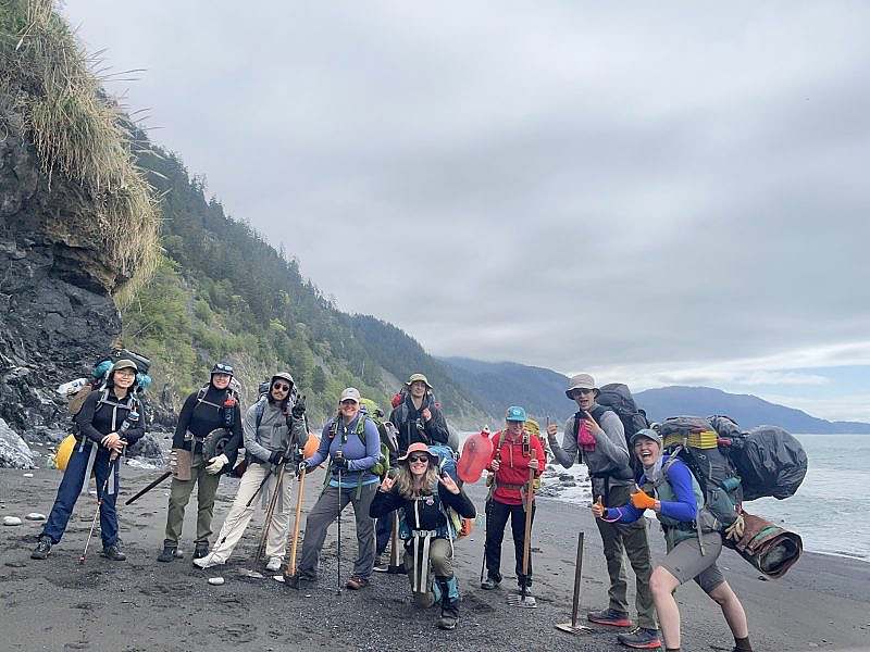 Nine people with large backpacks and trailbuilding tools smile along a gray sand beach with the ocean and mountain in the background.