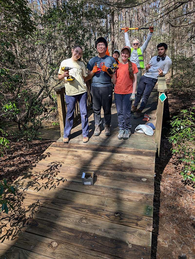 A group of 6 students pose on the wooden bridge they had just repaired. Wearing work clothes, gloves, and posing with power tools.
