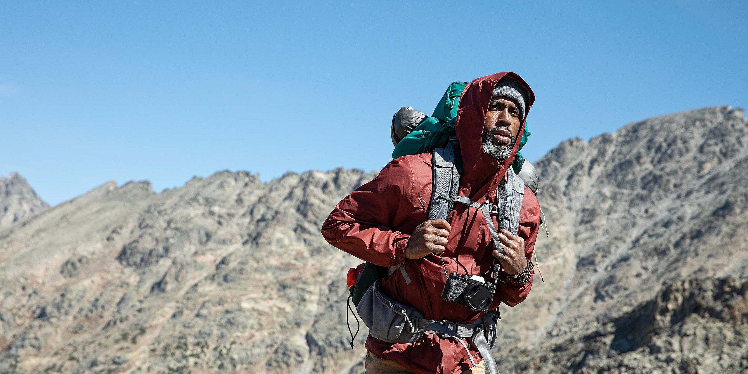 Hiker holds their pack straps as they hike with a warm hat and jacket with a hood with a blue sky and rugged ridge in the background.