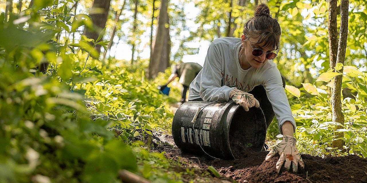 Volunteer in work gloves scoops soil into a black five-gallon bucket.
